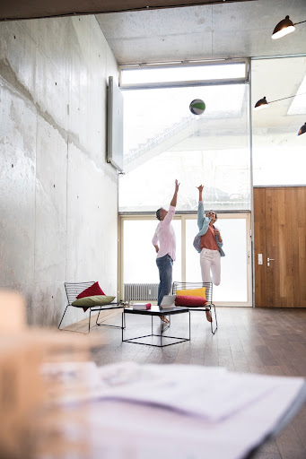 Two people in a modern concrete office space throwing a ball into the air. There are 2 chairs and a table with an out-of-focus desk in the foreground.