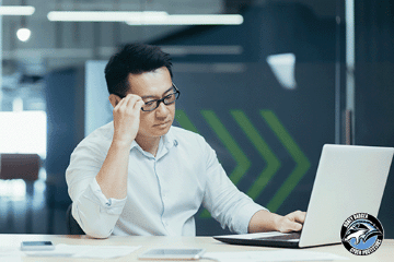 A man is sitting at a desk in an office space and holding his hand to his head while looking at a laptop computer.